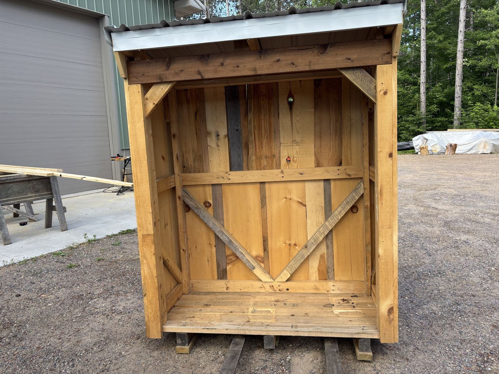 Wide open-front firewood shed with a gray steel roof and V-braced back wall