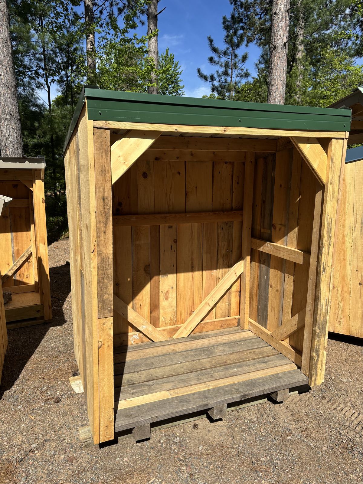 Open-front wood shed with a green steel roof, standing on gravel among tall pines